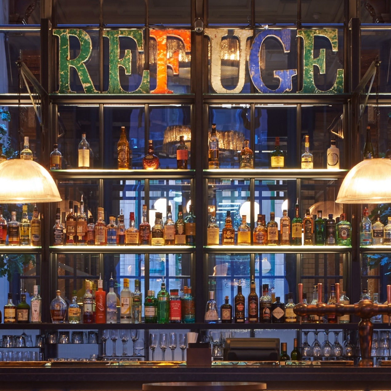 A bar with two art deco lamps hanging from the ceiling above with lots of bottles on the shelves with coloured letters spelling out REFUGE at the top